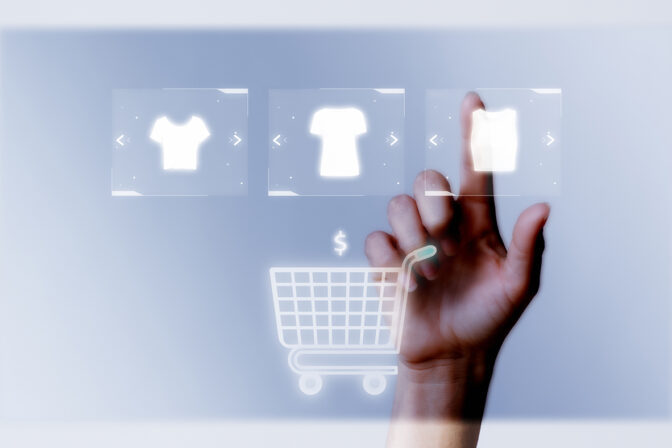 A human hand reaches up to touch a touch screen with three pieces of apparel on it. There is also an image of a shopping cart on the touch screen.