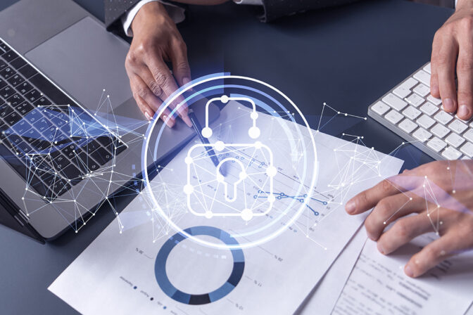 The image shows a pair of hands over a modern workspace. The left hand is using a pen on a pad of paper showing graphical interface elements, possibly for security management, while the right hand is typing on a keyboard. There are overlay graphics suggesting digital security concepts, such as a padlock icon connected with nodes and lines, symbolizing network security. Underneath the hands are printed materials that seem to be related to data or reports. The image conveys a concept of cybersecurity best practices or security information management in a corporate setting.