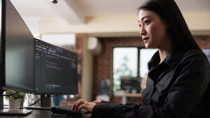 The image shows an Asian woman focused on her work at a modern, multi-monitor computer workstation. She is wearing a dark blouse, and her workspace is well-lit with natural light, suggesting a comfortable and contemporary office environment. Her posture and the concentration on her face indicate she is deeply engaged in a task, possibly related to cybersecurity, given the content on the screens.