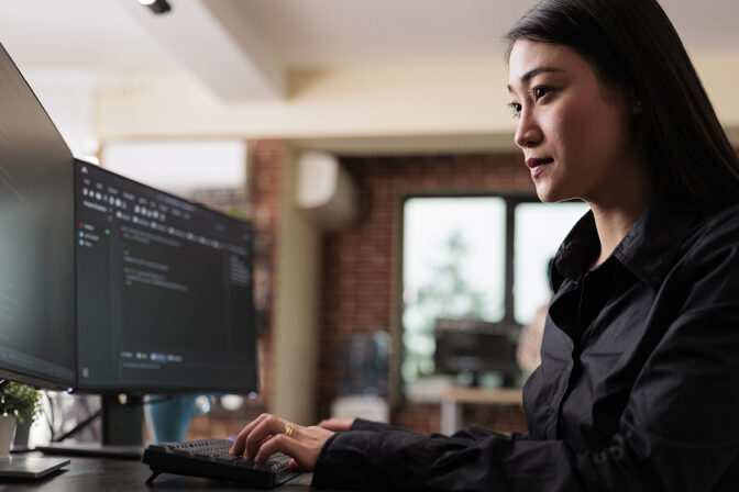 The image shows an Asian woman focused on her work at a modern, multi-monitor computer workstation. She is wearing a dark blouse, and her workspace is well-lit with natural light, suggesting a comfortable and contemporary office environment. Her posture and the concentration on her face indicate she is deeply engaged in a task, possibly related to cybersecurity, given the content on the screens.