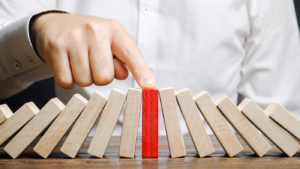 The image shows a close-up of a person's hand pushing a red domino, which stands out among a sequence of white dominoes set on a wooden surface. The action captures the moment before the red domino is likely to fall and initiate a chain reaction with the other dominoes. The person is wearing a white shirt, and only their hand is visible. The background is neutral and out of focus, emphasizing the dominoes and the action.