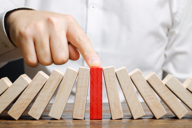 The image shows a close-up of a person's hand pushing a red domino, which stands out among a sequence of white dominoes set on a wooden surface. The action captures the moment before the red domino is likely to fall and initiate a chain reaction with the other dominoes. The person is wearing a white shirt, and only their hand is visible. The background is neutral and out of focus, emphasizing the dominoes and the action.