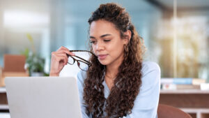 The image depicts a woman sitting in front of a laptop. She appears focused and contemplative, holding a pair of glasses in her hand while looking at the screen. Her hair is styled in long, curly locks that cascade down her shoulders. She wears a light blue blouse, which suggests a professional or business casual environment. The setting appears to be a modern, well-lit office with a blurred background, indicating a possible workspace.