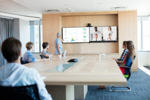 A modern conference room equipped with the latest technology trends for efficient virtual meetings, featuring a large screen displaying data charts and connecting remote participants. The room is well-lit with natural light, and attendees are seated at a sleek wooden table, focusing attentively on the presenter and the virtual participants on the screen.