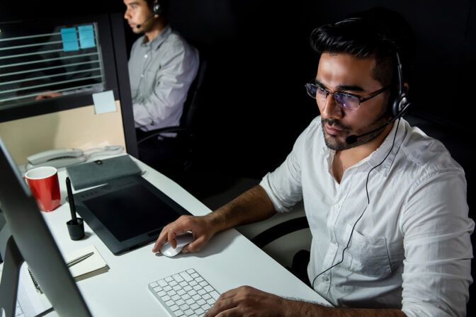 A man wearing a headset with a microphone sits at a managed IT help desk in a dimly lit office environment. He is focused on a computer monitor, operating a mouse and keyboard, with a digital drawing tablet, a red cup, and office supplies on the desk. Another person with a headset can be seen in the background, suggesting a collaborative workplace.