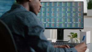 A professional working on cybersecurity in the financial sector, following SEC cybersecurity guidance. The person is seated in front of a large monitor displaying a complex array of stock market analytics and data encryption measures. This workspace exemplifies a secure environment for monitoring and protecting sensitive financial data against cyber threats, in compliance with regulatory standards for security and confidentiality in the stock exchange market.