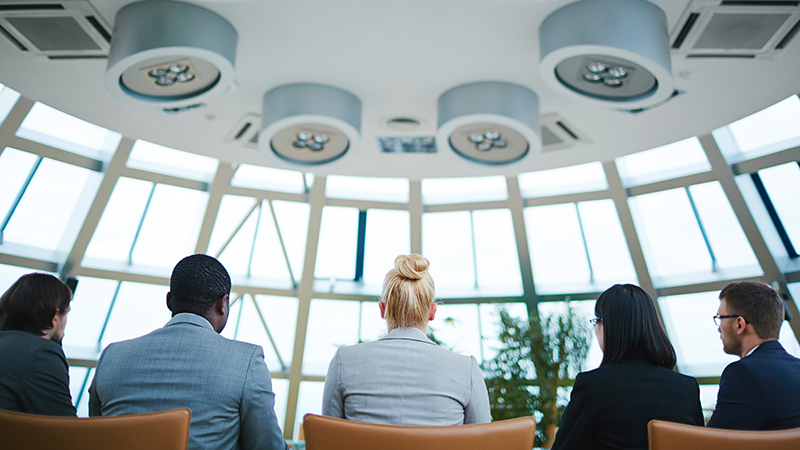 Five people sitting in a conference room with a modern, circular ceiling design featuring integrated lighting and ventilation systems. They are facing large windows that provide a view of the outside.