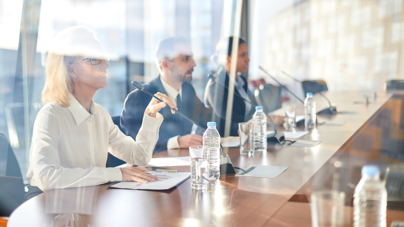 Three panelists, including an older woman in glasses and two men, speaking into microphones at a conference table with water bottles and documents in front of them. They are in a modern, glass-enclosed room.