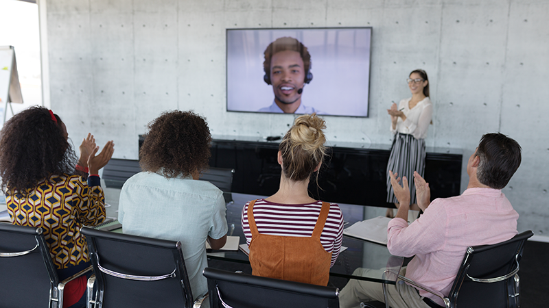 A diverse group of people sitting in a conference room, watching a man on a large video screen who is speaking via video conference. The group is clapping, and a woman stands near the screen, smiling and gesturing.