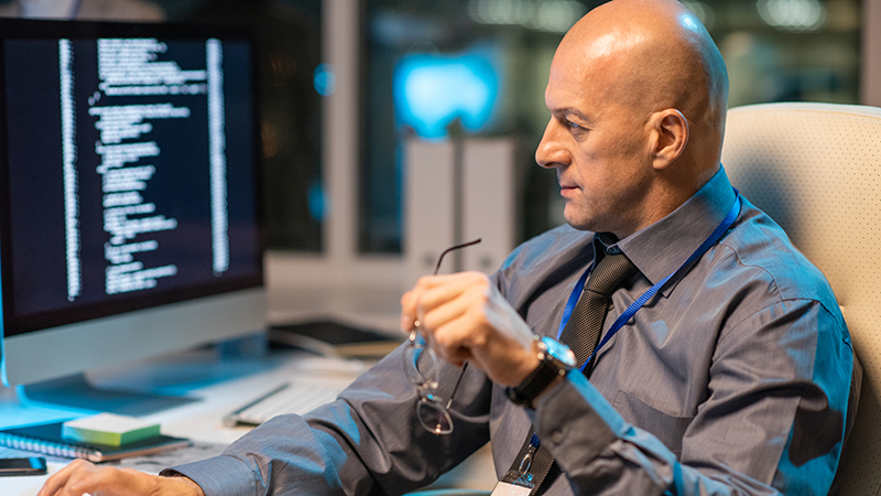 The image depicts a middle-aged, bald man in a blue shirt and tie, seated at a desk. He is holding his glasses in one hand and appears to be focused on the code displayed on a computer screen in front of him. The setting suggests a modern office environment, possibly at night given the lighting, and his lanyard implies he may be a virtual security engineer.