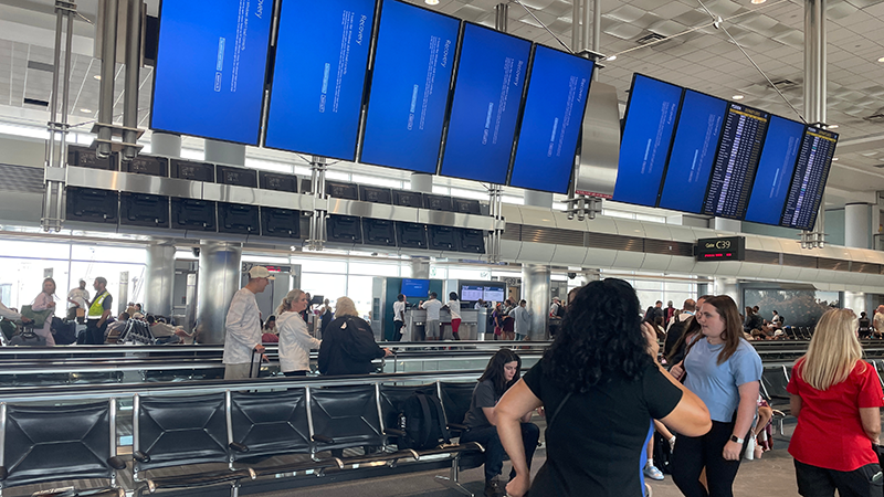 Screens showing the Microsoft "Blue Screen of Death" at the Denver airport as passengers appear upset in the foreground and background as they try and find information on the CrowdStrike incident.