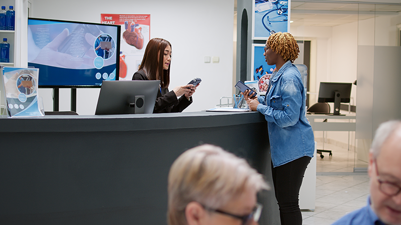 A woman with a check-in device behind a counter, and a woman with her phone checking in for an appointment. The facility is a healthcare office.