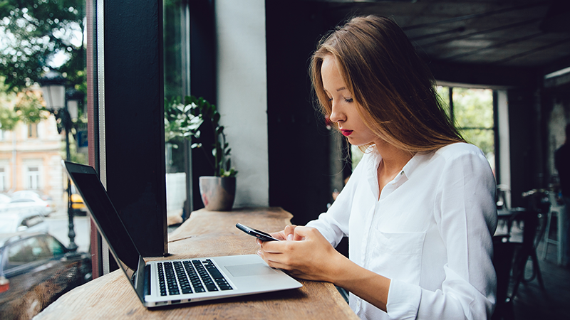 A woman site at a cafe using her phone and a laptop indicating that BYOD security is essential.