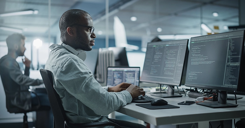 A cybersecurity professional conducts a penetration test on multiple monitors, analyzing code and system vulnerabilities. Another individual is visible in the background working on a similar setup, representing a collaborative environment for vulnerability scanning and pen testing.