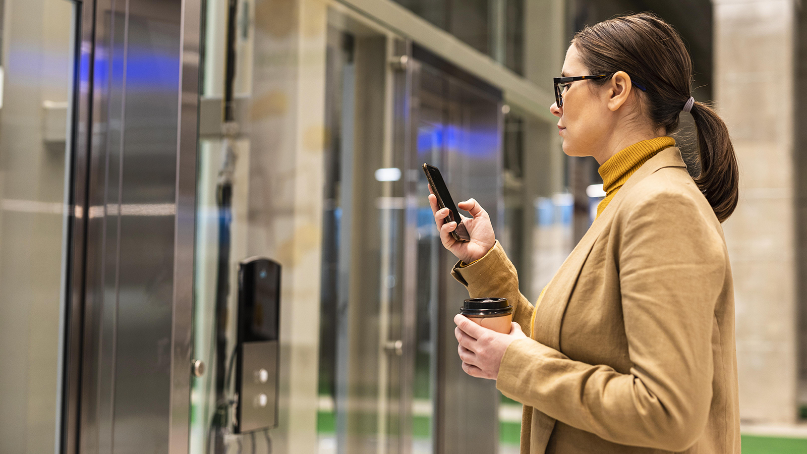 Woman using smartphone to access a secure glass door with an access control panel, holding a coffee cup in her other hand, in a modern office setting.