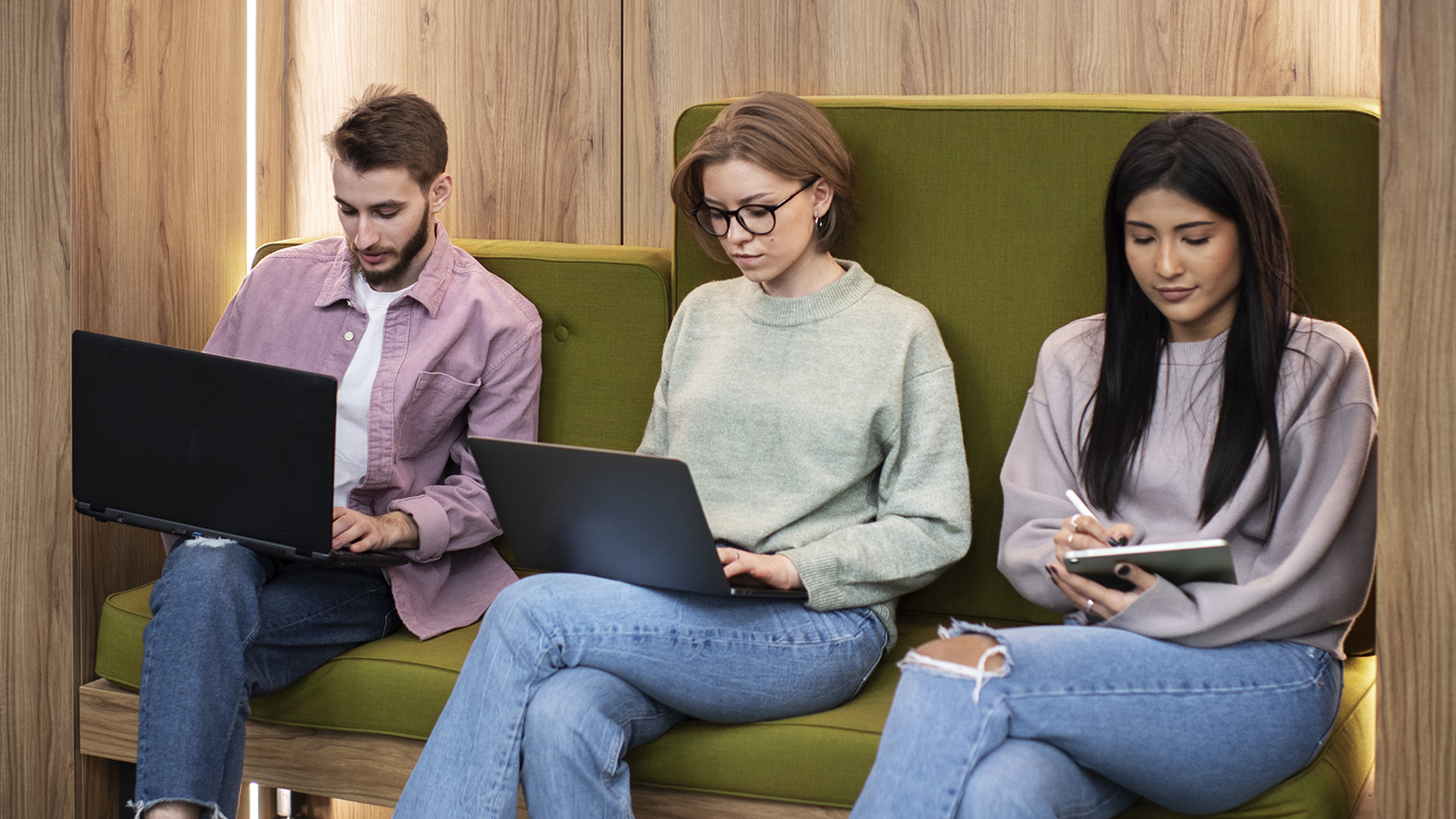 Three young professionals sitting together on a green cushioned bench in a modern, wood-paneled workspace. Two of them are working on laptops, and one is taking notes on a tablet. They are focused and engaged, collaborating in a casual, relaxed setting.