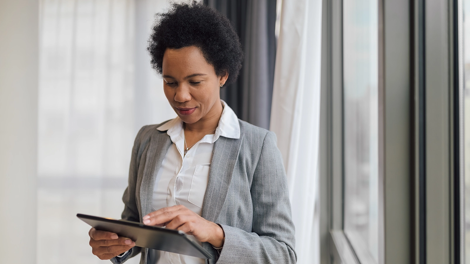 A professional woman in a gray suit stands near a window, engaging with a tablet, highlighting the importance of mobile device security in workplace environments.