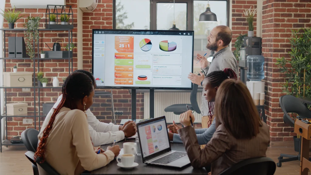 A group of diverse professionals in a modern office with exposed brick walls participating in an IT strategy session. A bearded man stands at the front, presenting business analytics on a large digital screen displaying charts, graphs, and data insights. Three colleagues sit around a table, taking notes and working on laptops, engaging in the discussion. The office features shelves with plants, a water cooler, and industrial-style lighting.