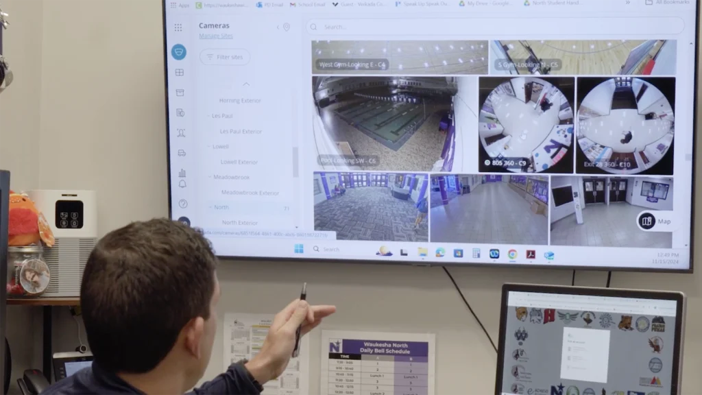 A security personnel member points at a large wall-mounted screen displaying a Verkada security camera dashboard in a school security office. The dashboard shows multiple live camera feeds, including gymnasiums, hallways, and entrances. A computer monitor below also displays login options. A school bell schedule for Waukesha North is posted on the wall, indicating the location is part of the School District of Waukesha's security operations.