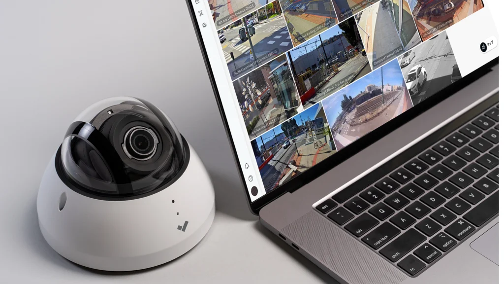 A Verkada dome security camera placed next to a MacBook laptop displaying a video surveillance dashboard with multiple live security camera feeds from different locations. The camera has a sleek white and black design, and the laptop screen shows a modern user interface for monitoring footage.