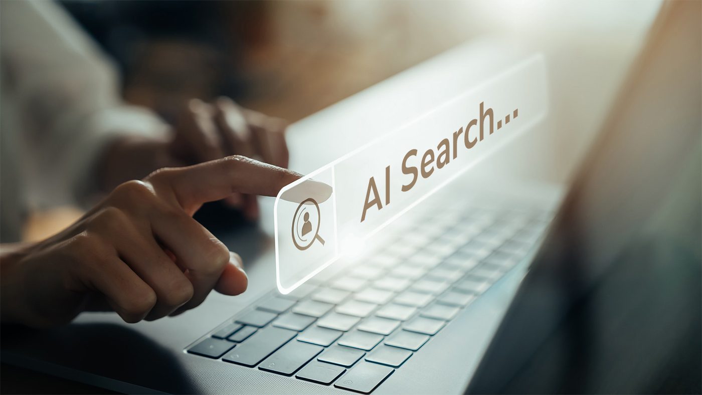 Realistic image of a person’s hand resting on a laptop keyboard with one finger of another hand approaching a search bar with the words “AI Search.”