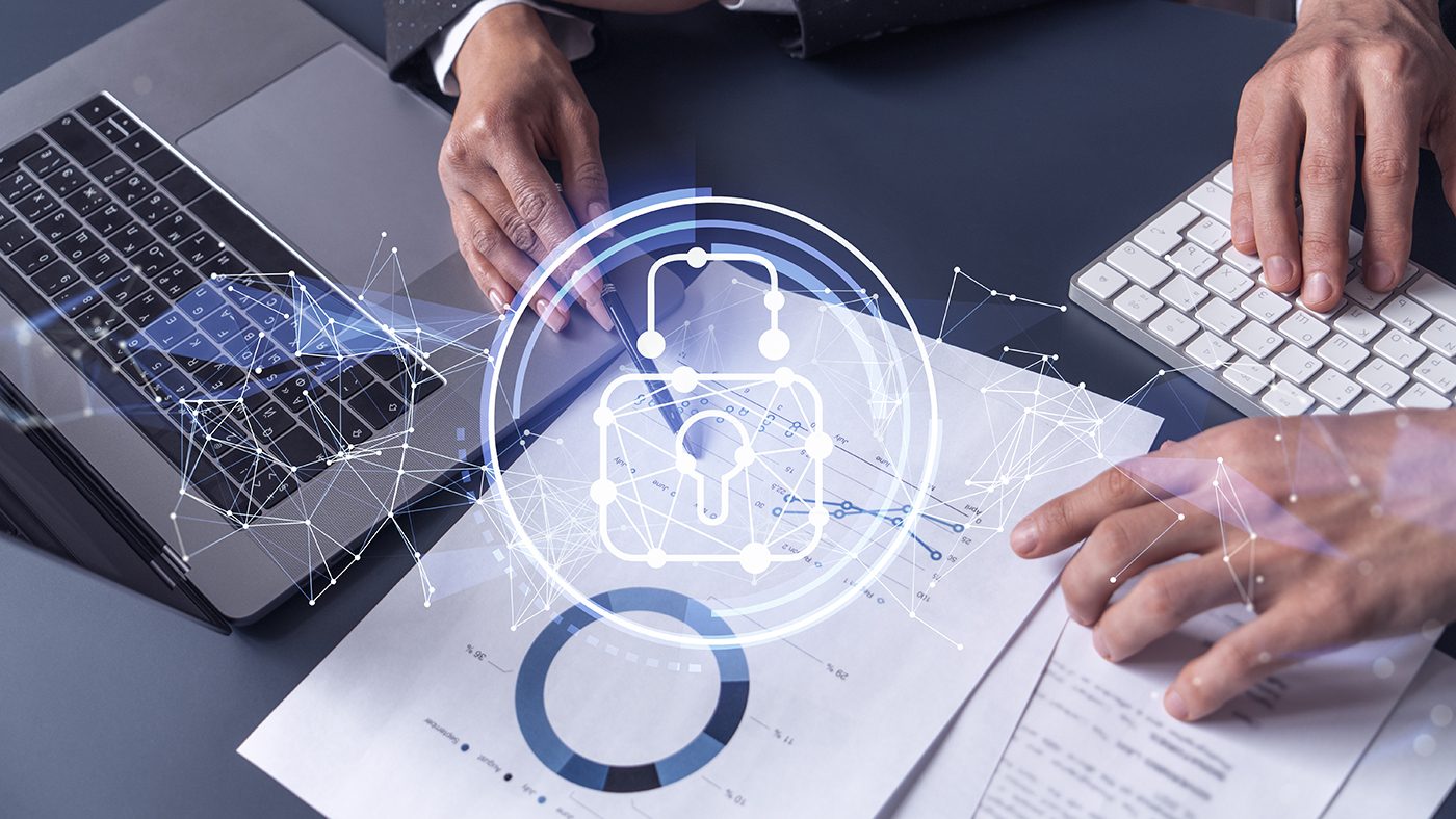 The image shows a pair of hands over a modern workspace. The left hand is using a pen on a pad of paper showing graphical interface elements, possibly for security management, while the right hand is typing on a keyboard. There are overlay graphics suggesting digital security concepts, such as a padlock icon connected with nodes and lines, symbolizing network security. Underneath the hands are printed materials that seem to be related to data or reports. The image conveys a concept of cybersecurity best practices or security information management in a corporate setting.
