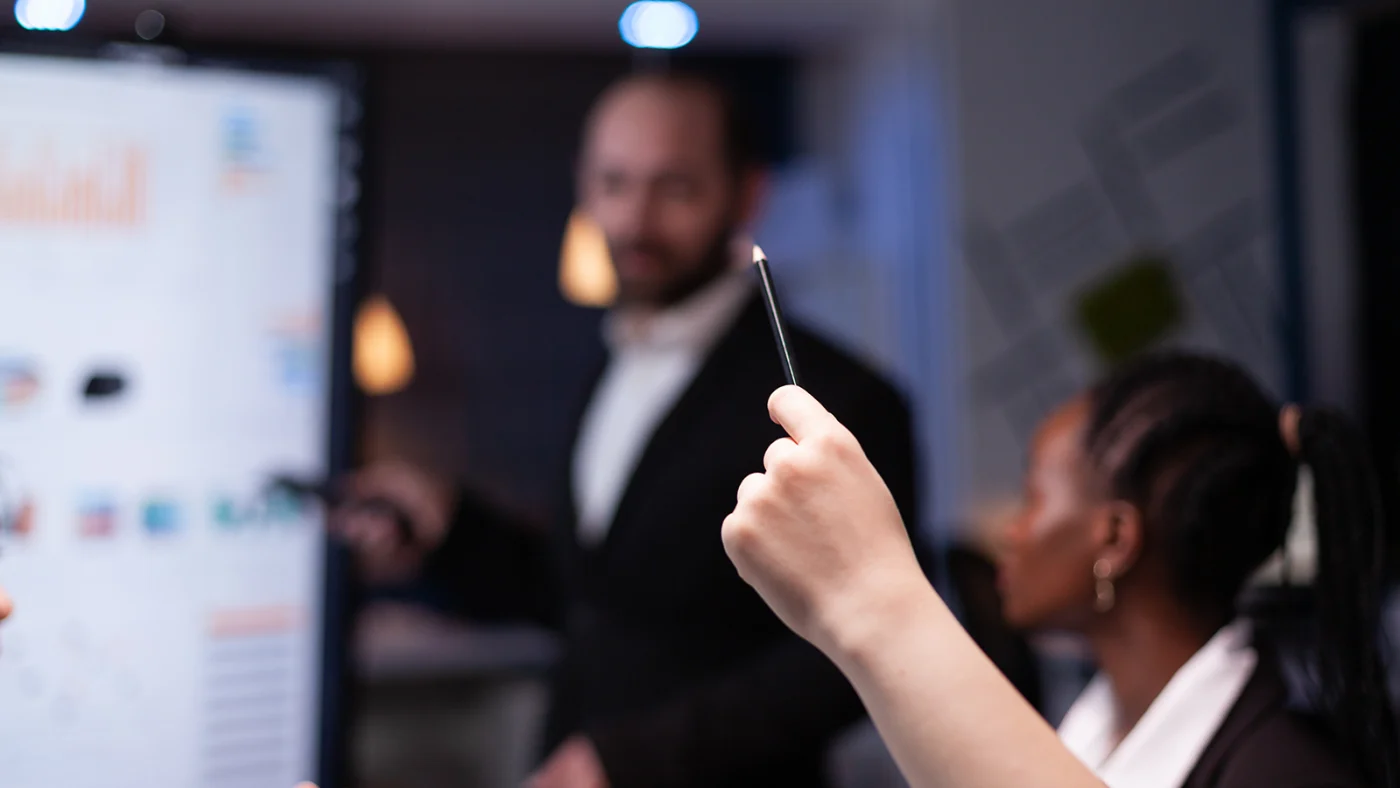 A close-up of a participant raising a pen during the Microsoft Power Platform Envisioning Workshop. In the blurred background, a presenter points to a digital screen displaying charts, engaging with the group in a collaborative discussion.