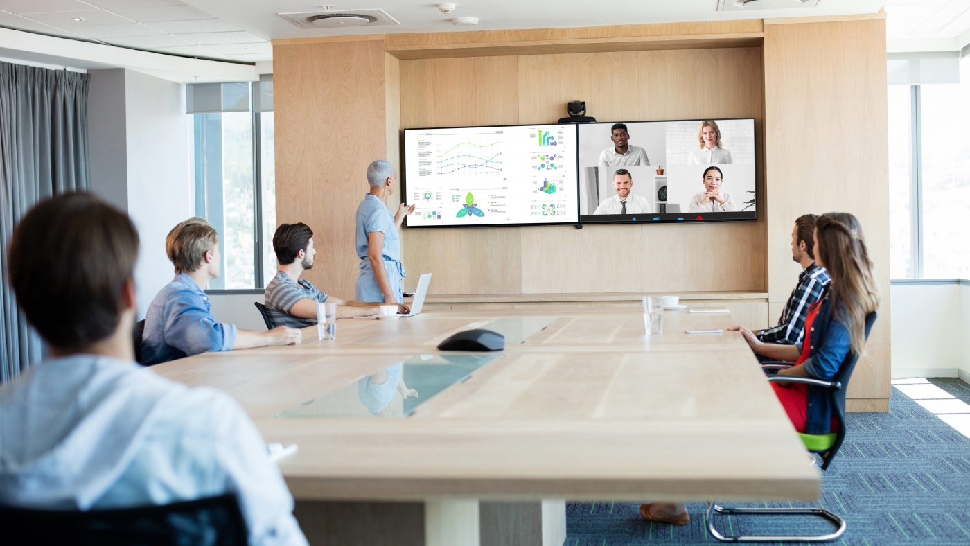 A modern conference room equipped with the latest technology trends for efficient virtual meetings, featuring a large screen displaying data charts and connecting remote participants. The room is well-lit with natural light, and attendees are seated at a sleek wooden table, focusing attentively on the presenter and the virtual participants on the screen.