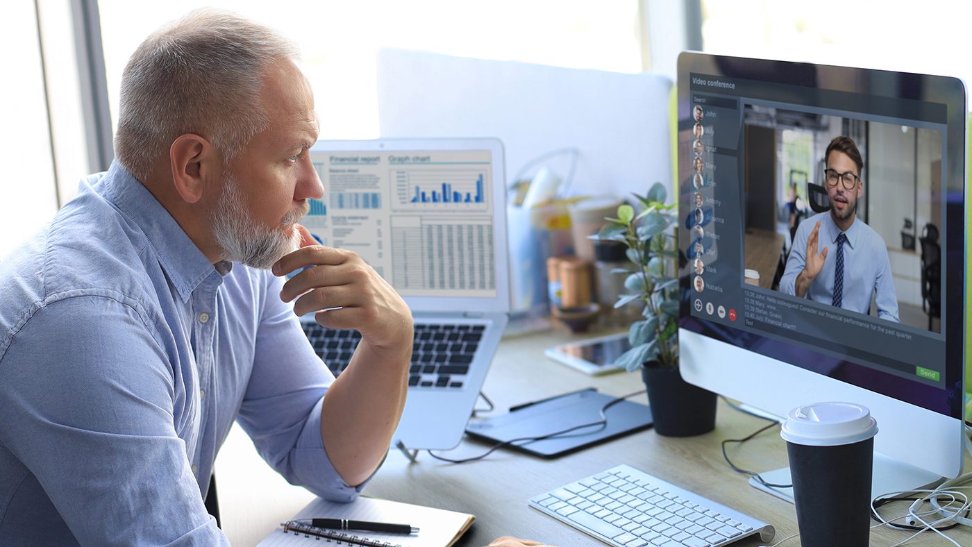 A mature businessman with a thoughtful expression is participating in a video conference on his desktop computer. The screen shows a younger man gesturing as if explaining something. The businessman's desk features an open laptop displaying financial charts, a notepad, and a closed cup of coffee, indicating a professional setting. This image reflects how businesses might utilize virtual Chief Information Officer (vCIO) services to receive remote, strategic consultation on IT matters, exemplifying why to use a vCIO for expertise without the need for an in-house executive.